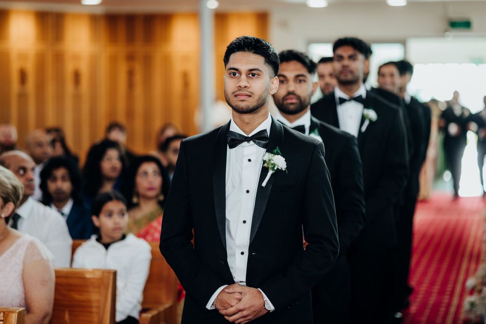 Groom in black tuxedo with boutonniere stands at altar during wedding ceremony with groomsmen behind him.