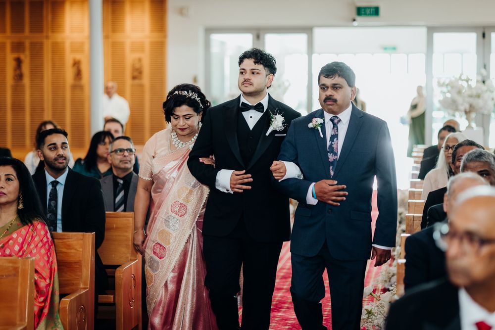 Groom in tuxedo walks down church aisle escorted by two family members during wedding ceremony.