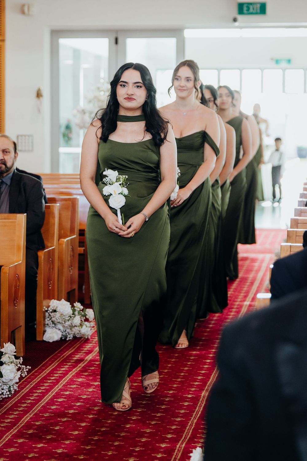 Bridesmaids in olive green gowns walk down a red-carpeted church aisle carrying white flowers.