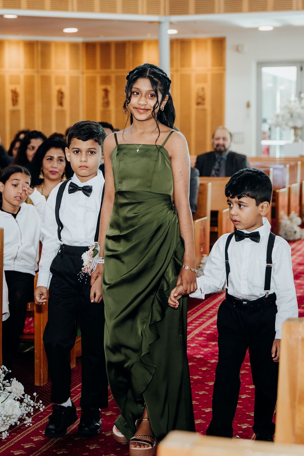 Young bridesmaid in green satin gown walks down church aisle with young boys in suspenders and bow ties.