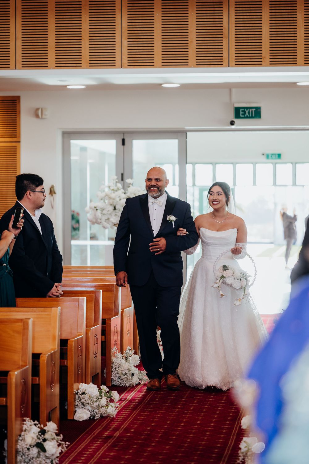 Bride walks down the church aisle with escort, smiling joyfully in a white strapless gown during wedding ceremony.