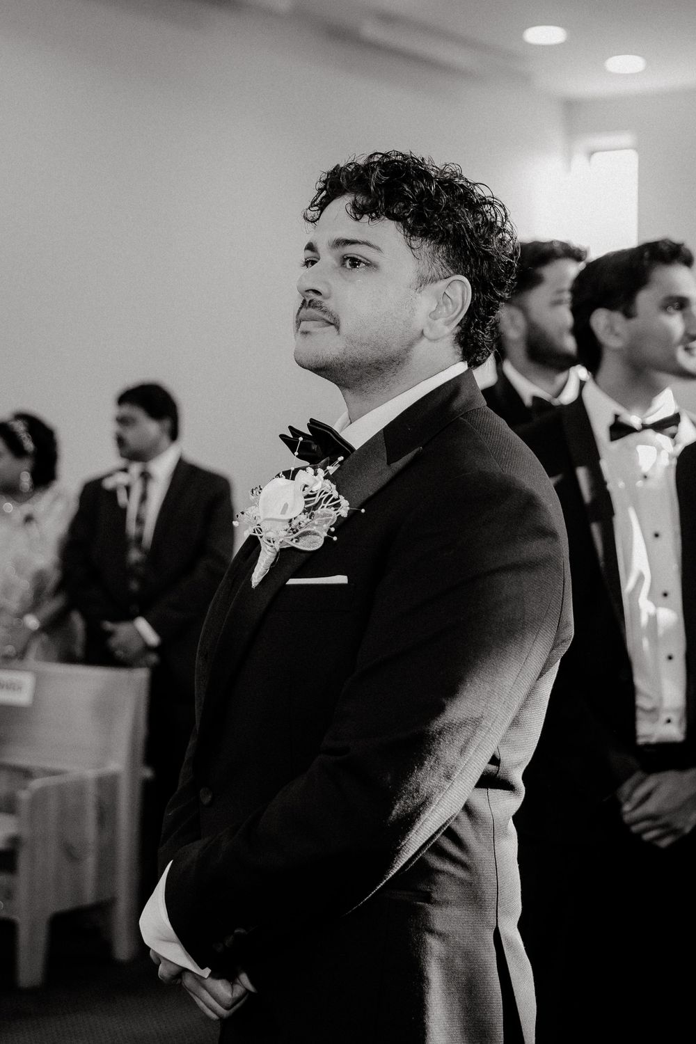 Groom in tuxedo with boutonniere watching bride walk down aisle at wedding ceremony.