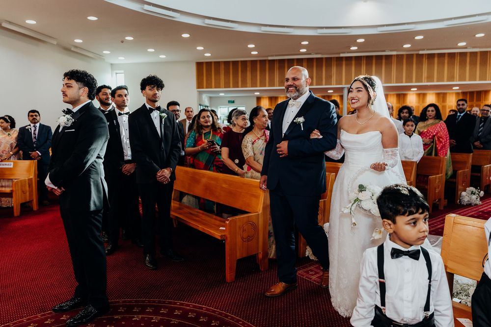 Bride walks down church aisle with escort during wedding ceremony, guests seated in pews watching the procession.