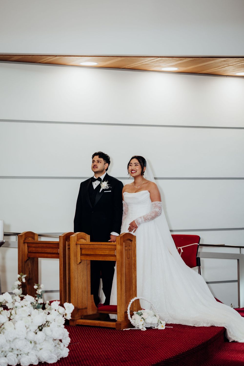Bride and groom standing at church altar during wedding ceremony with white floral arrangements on red carpet steps.