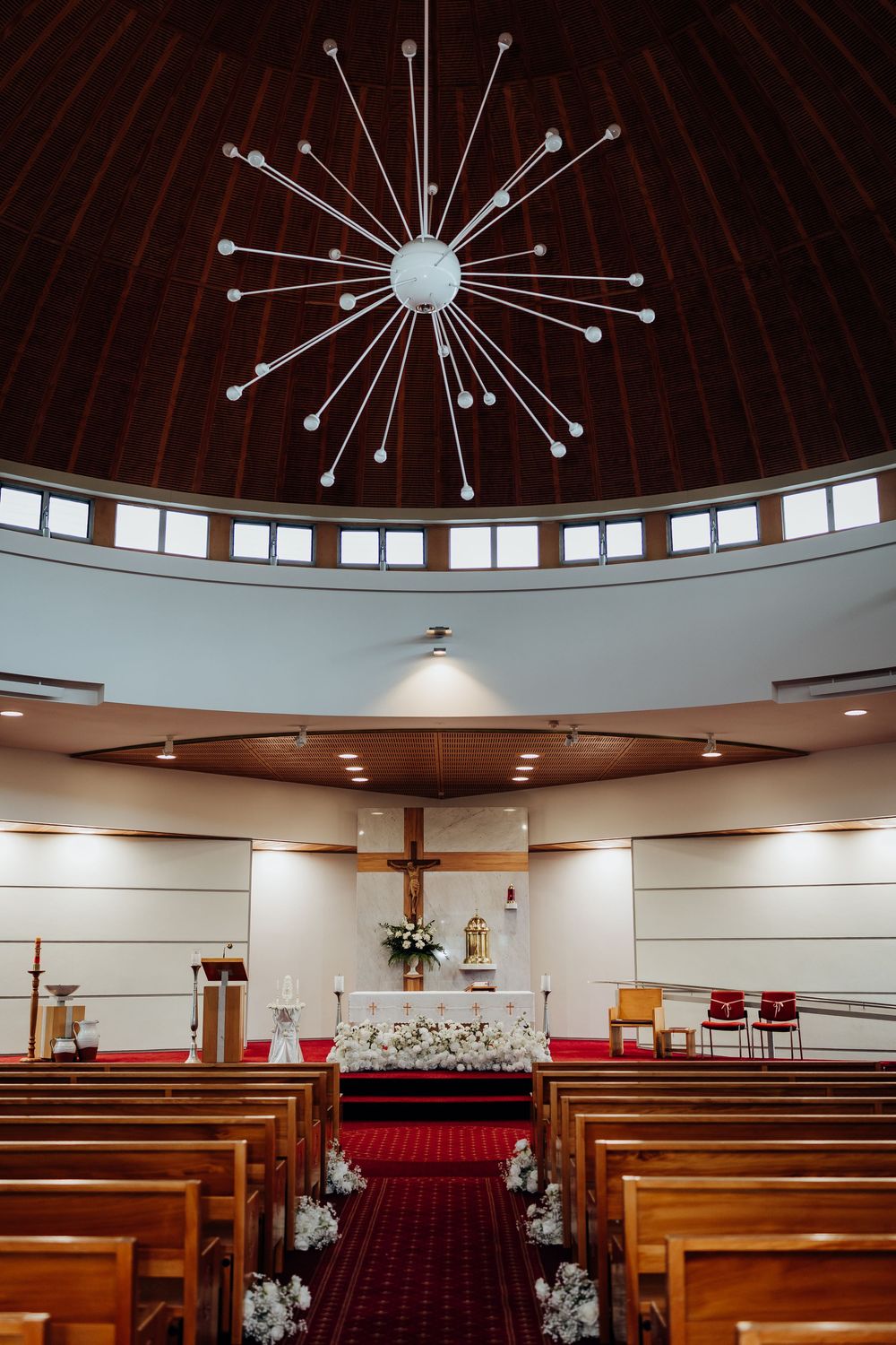 Elegant church interior with domed ceiling, starburst chandelier, wooden pews, and floral-decorated altar for wedding ceremony.