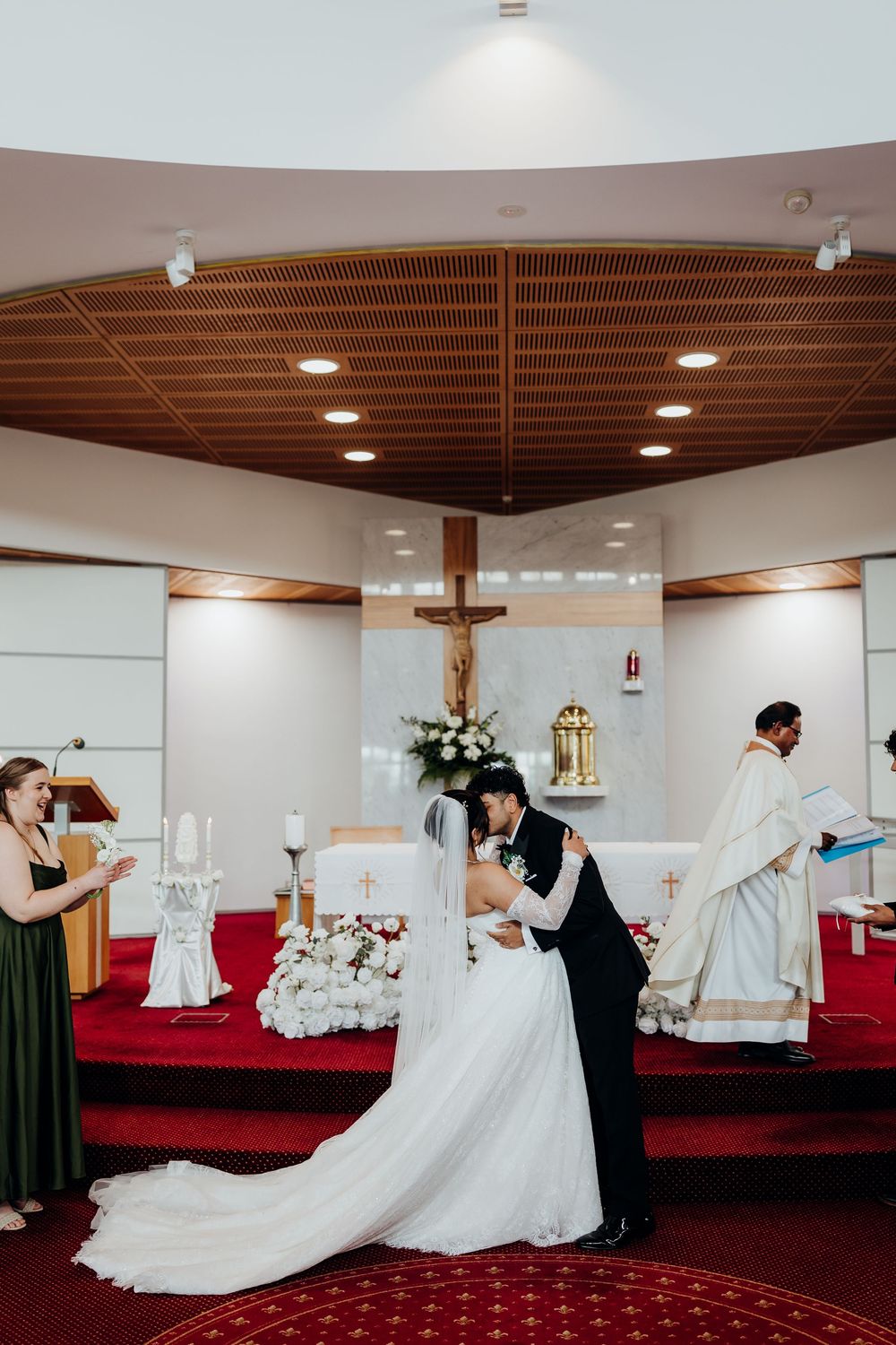 Bride and groom share first kiss at church altar during wedding ceremony with priest and bridesmaid nearby.