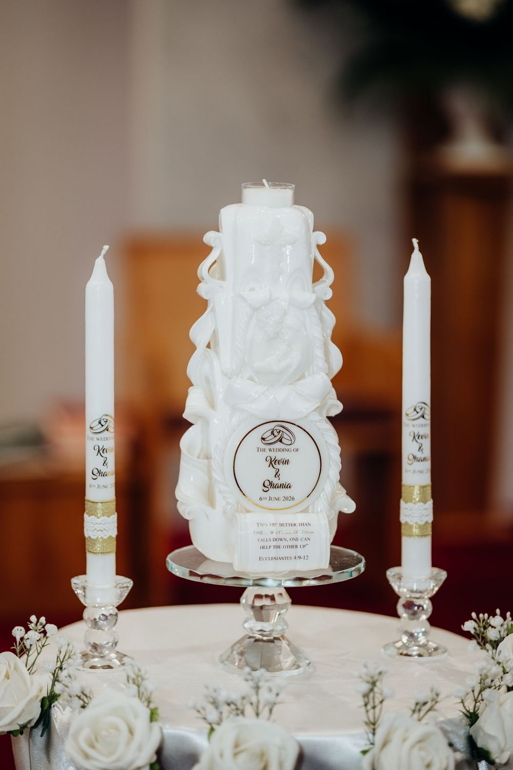 Elegant white unity candle set with carved details and personalized wedding card, flanked by taper candles and white roses.