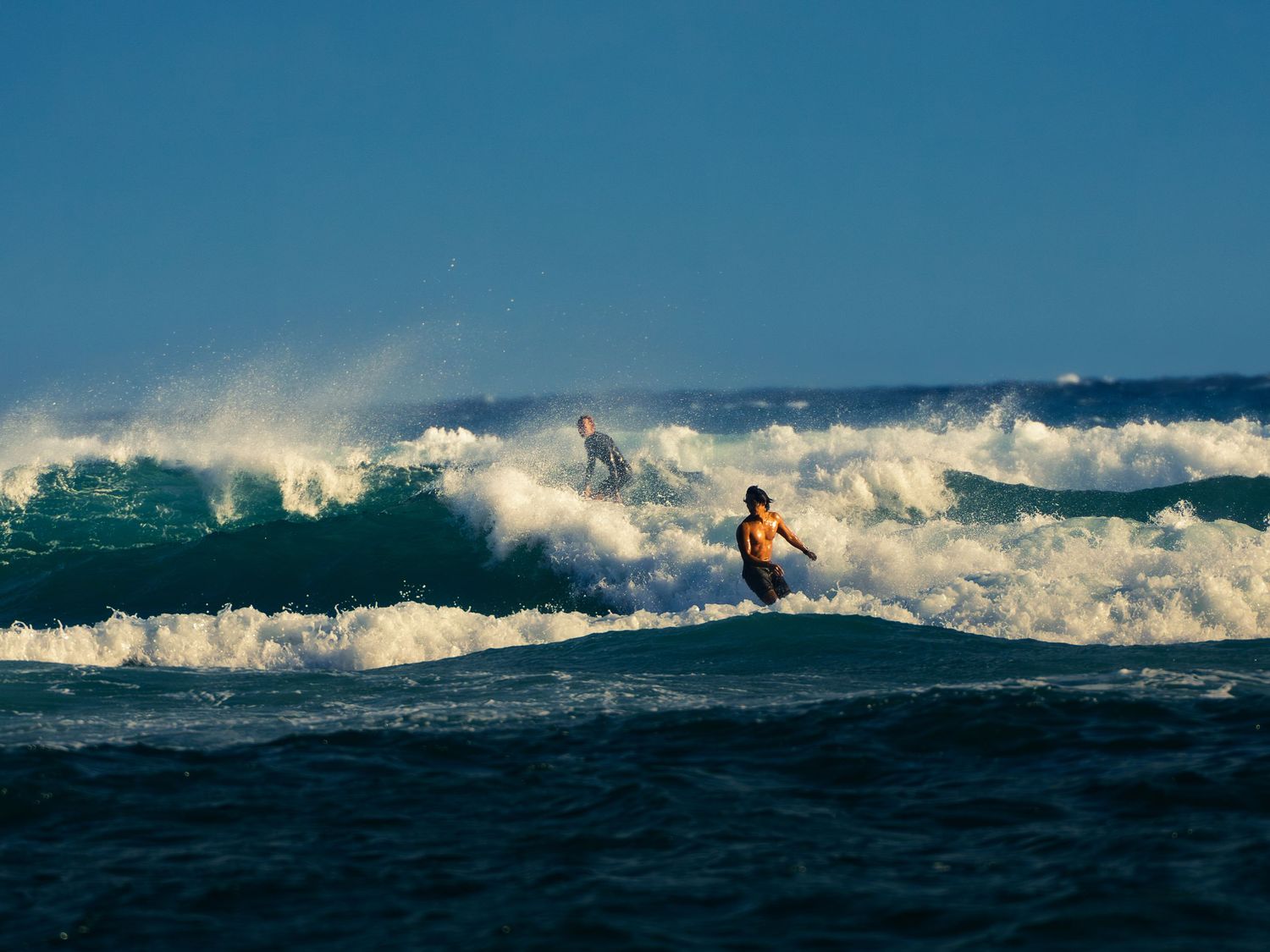 A shirtless surfer in dark boardshorts rides a breaking wave in warm light off Waikiki, a second surfer in the whitewater behind him.