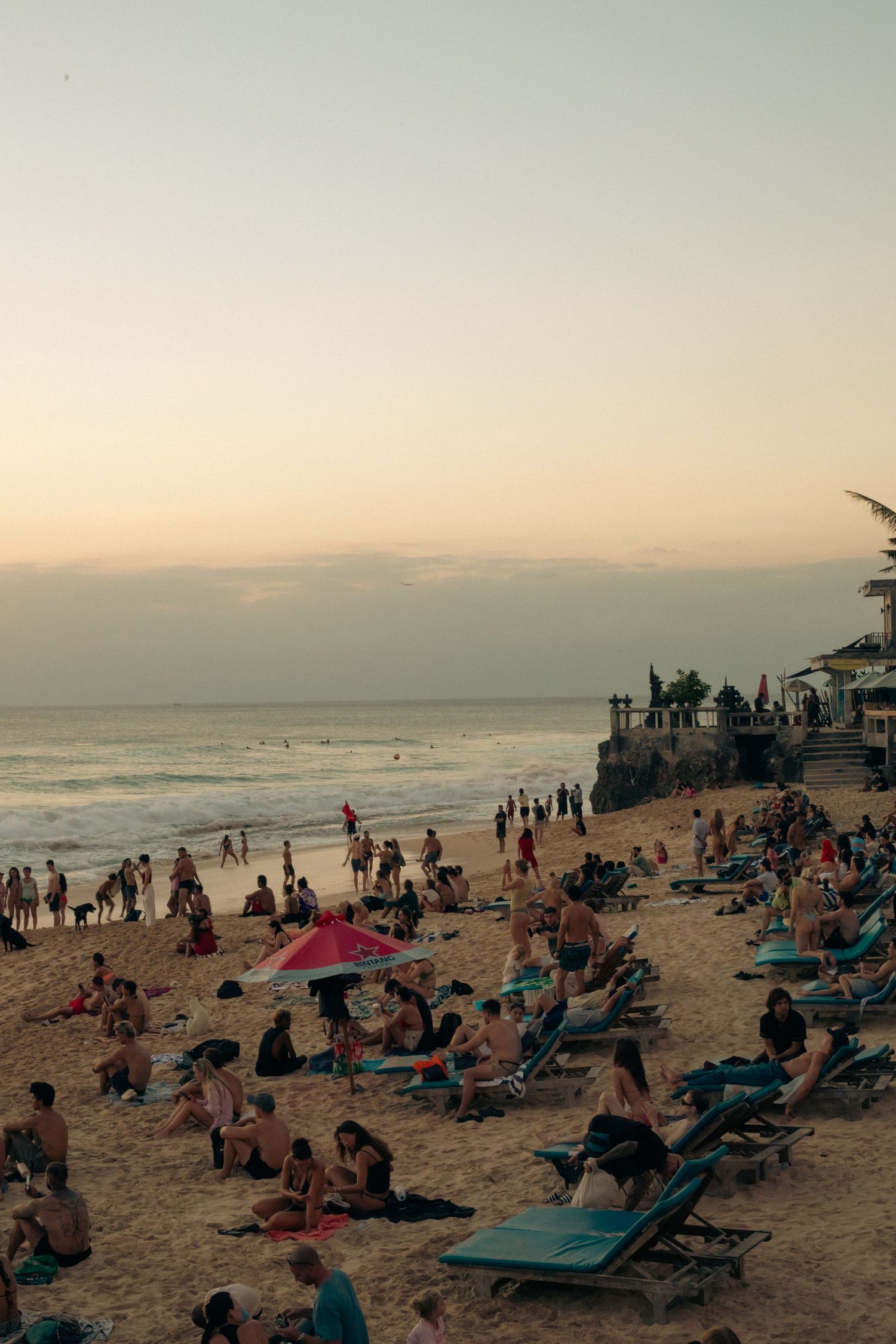 Crowded beach at sunset with tourists relaxing on lounge chairs and sand, ocean waves and a clifftop building in the background.
