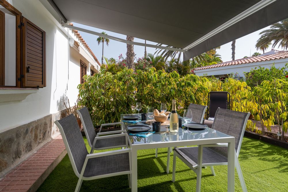 Outdoor dining area with gray chairs and glass table under a pergola, surrounded by lush green tropical garden.