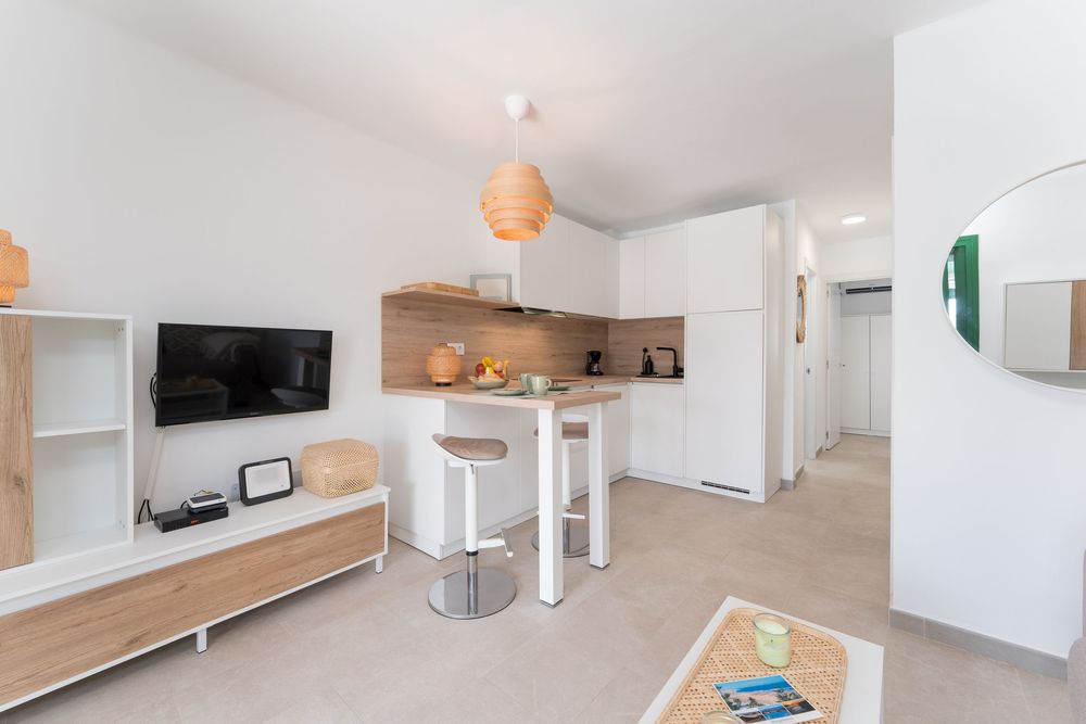 Modern studio apartment with open-plan kitchen, white cabinets, wooden accents, bar stools, and wall-mounted TV.