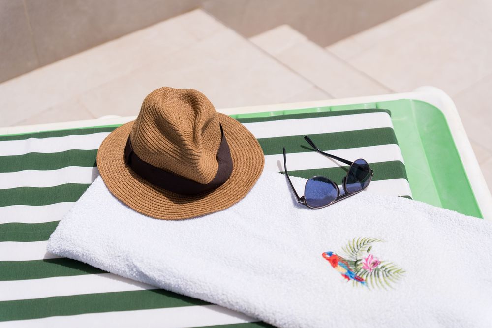 Straw hat and sunglasses on a striped green and white beach towel with parrot embroidery in sunny outdoor setting.