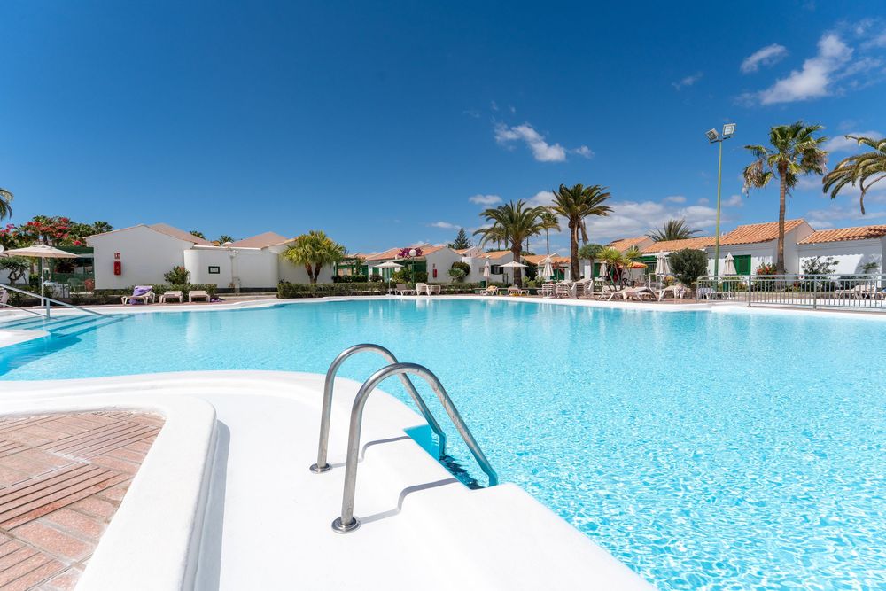 Large outdoor swimming pool with crystal-clear blue water, metal handrail, palm trees, and white resort villas under a sunny sky.