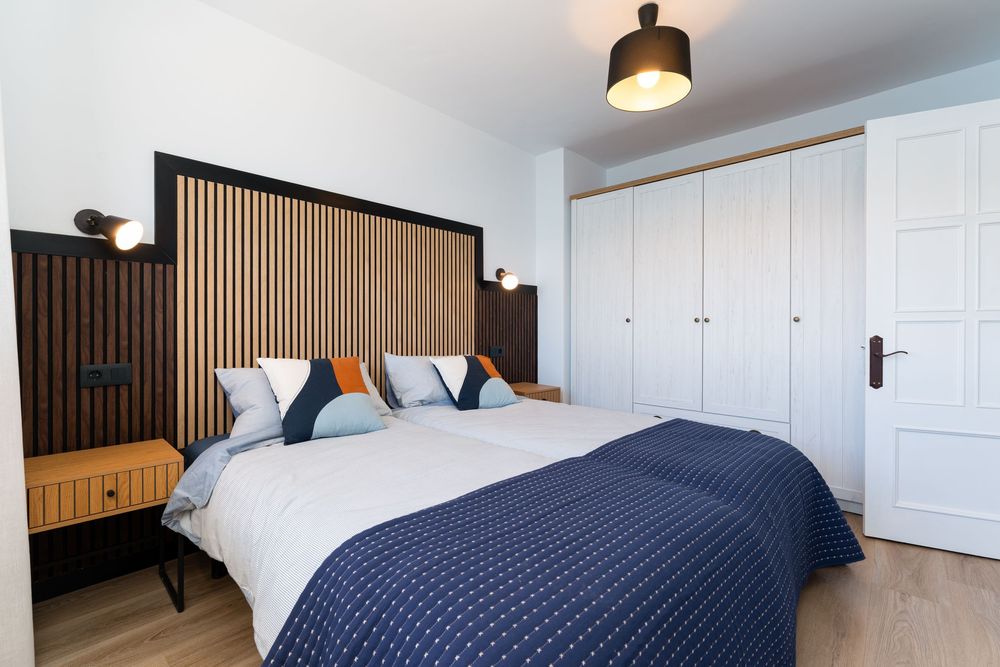Modern bedroom with navy blue bedding, wooden slatted headboard, white wardrobe, and orange accent pillows.