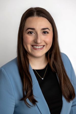 Professional headshot of a smiling young woman with long brown hair wearing a blue blazer and gold necklace.