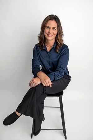 A professional woman in a navy blouse and black pants sits confidently on a stool against a white background.