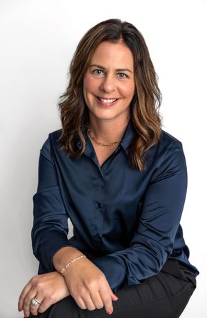 Professional woman in navy blue blouse smiling confidently, seated against white background, wearing gold necklace.