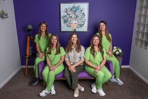 Five women pose in a purple room, four wearing green scrubs, one in casual wear, seated on a purple couch.