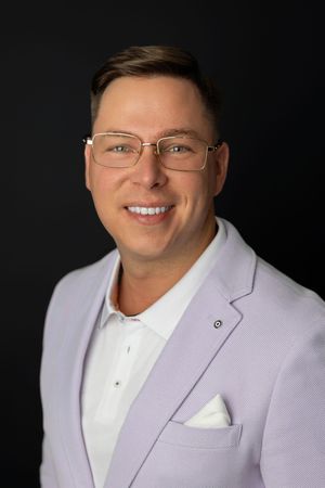 Smiling man in light purple blazer and glasses poses for professional headshot against dark background.