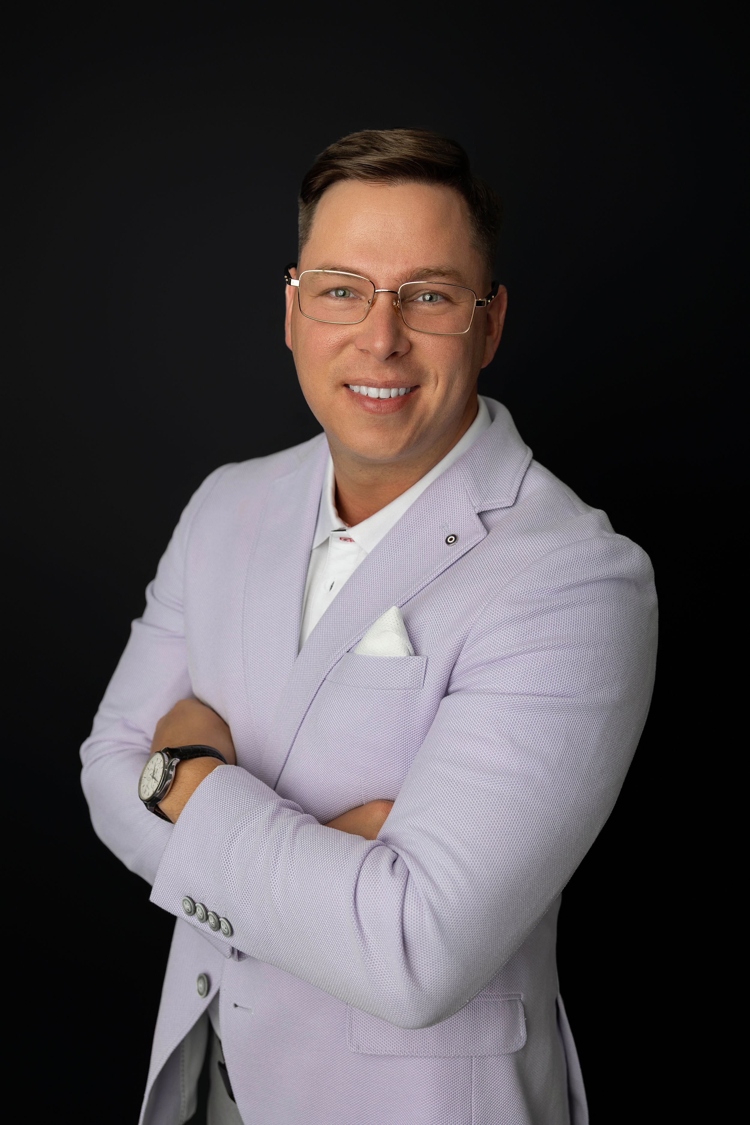 Professional man in lavender blazer and glasses smiling with arms crossed against dark background.