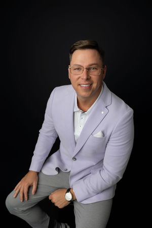 A smiling man in a lavender blazer and gray pants sits confidently against a dark background wearing glasses and a watch.