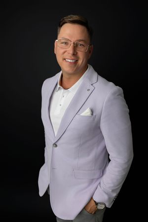 Smiling man in lavender blazer and white shirt with pocket square, wearing glasses, posing against dark background.
