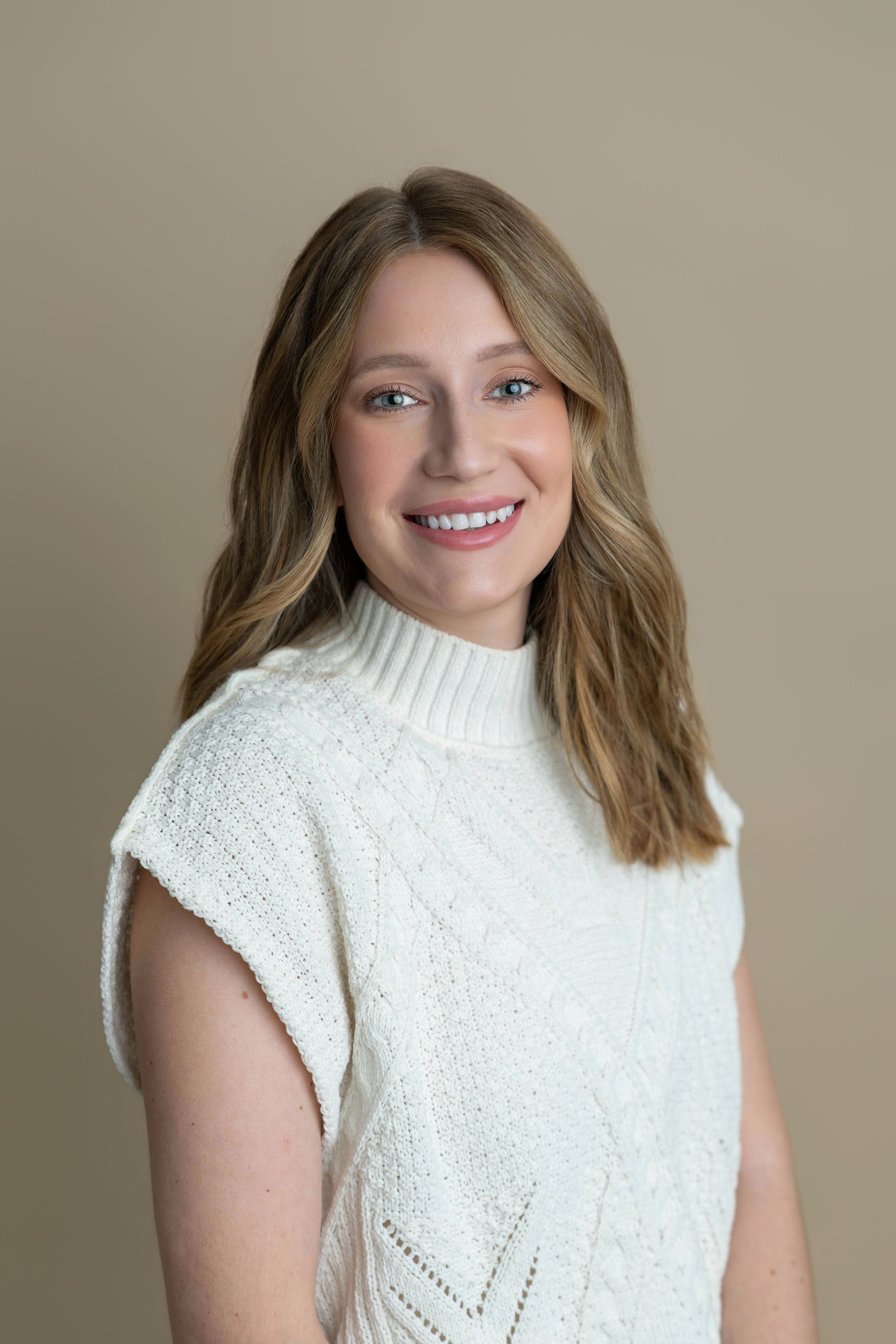 Smiling woman with blonde hair wearing a white cable-knit sleeveless sweater against a beige background.