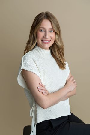 Smiling woman with blonde hair wearing a white sleeveless knit sweater, arms crossed, posed against a beige background.