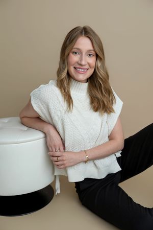 Smiling woman in white cable-knit vest and black pants sitting on floor leaning on white ottoman.