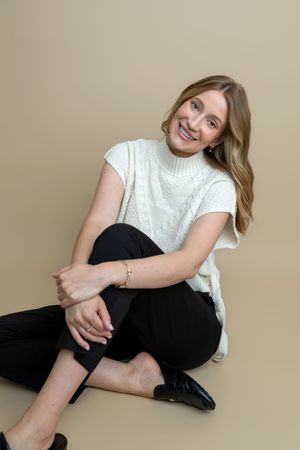 Smiling woman in white cable-knit sweater vest and black pants sitting on floor against beige background.