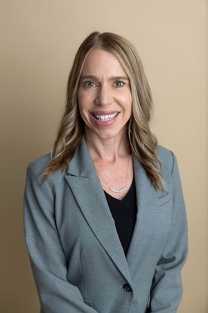 Professional woman in gray blazer smiling confidently against neutral background for business headshot.