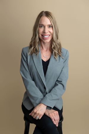 Smiling blonde woman in gray blazer and black top seated confidently against a neutral beige background.