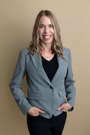 Smiling professional woman in gray blazer and black top poses confidently against a neutral beige background.