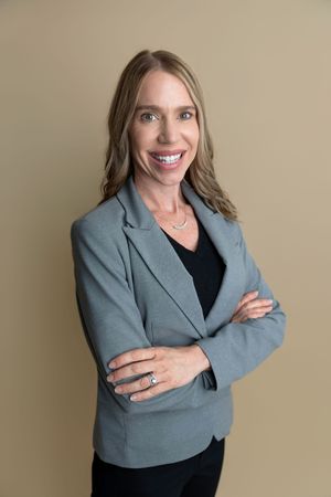 Professional woman in gray blazer smiling with arms crossed against beige background.