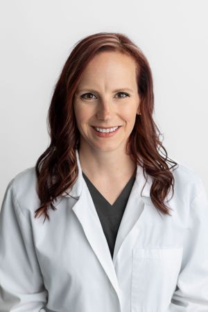 Female healthcare professional in white lab coat and dark scrubs smiling confidently against light background.