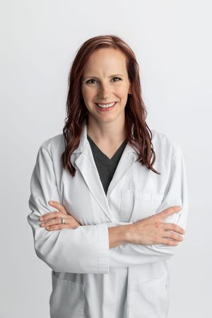 Smiling female doctor in white lab coat with arms crossed against light gray background.