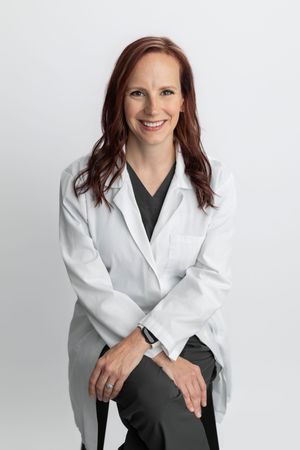 Smiling female doctor in white lab coat and dark scrubs sitting confidently against a light gray background.