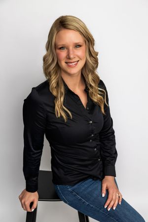 Smiling blonde woman in black satin shirt and jeans sitting on stool against white background.