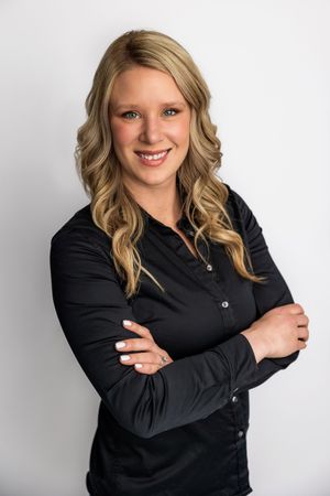 Smiling blonde woman in black blouse with arms crossed poses confidently against white background.