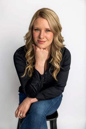 Smiling blonde woman in black shirt and jeans sitting on stool with hand on chin against white background.