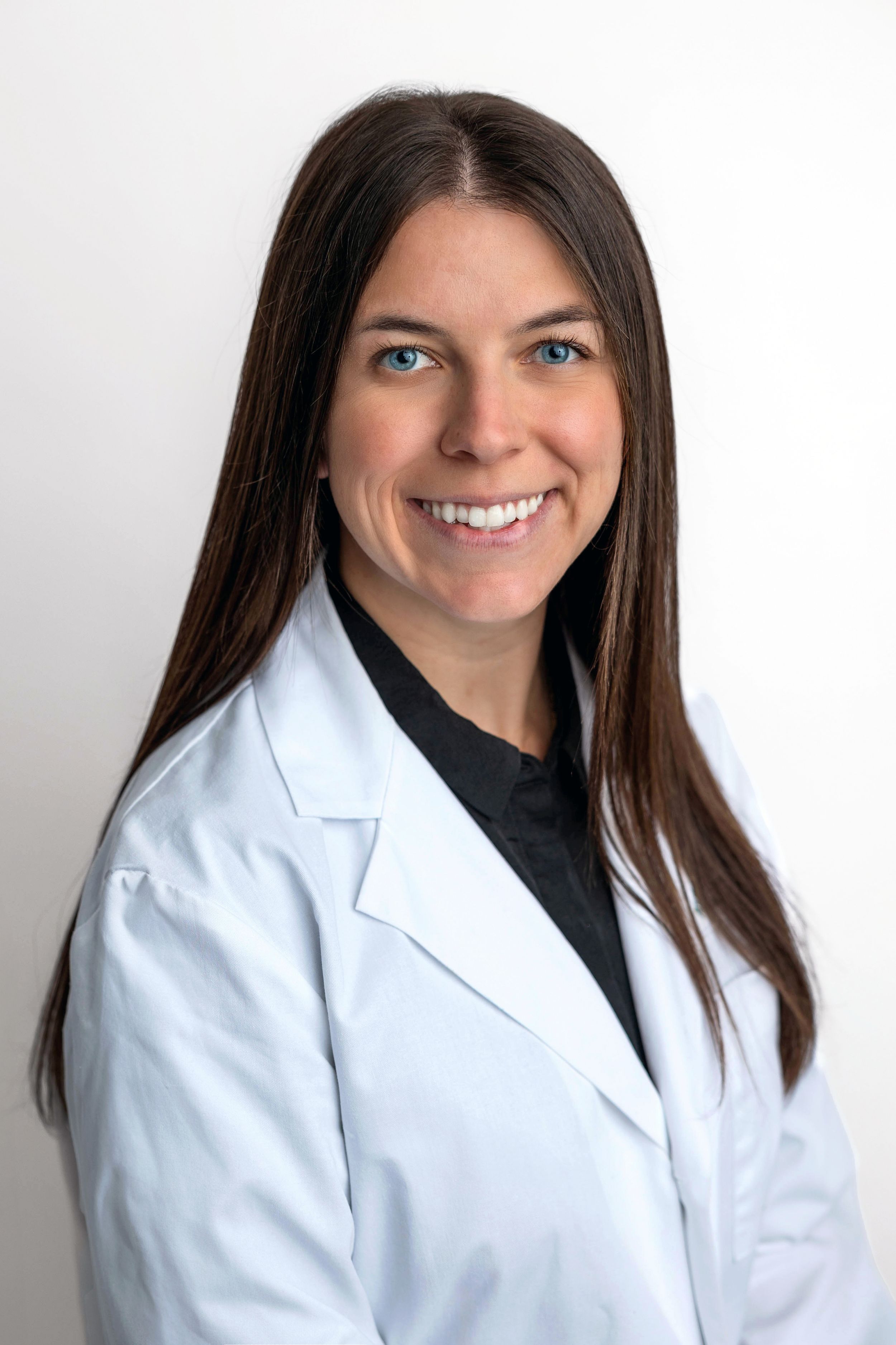 Smiling female healthcare professional with long brown hair wearing a white lab coat and black shirt against a white background.