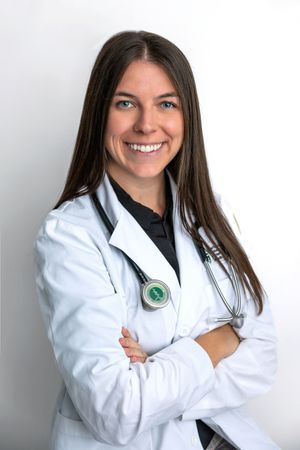 Smiling female doctor in white lab coat with stethoscope, arms crossed, posing confidently against white background.