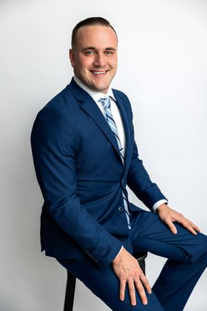 Professional man in navy blue suit and striped tie sitting confidently on stool against white background.