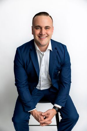 Smiling man in a navy blue suit sitting on a stool against a white background.