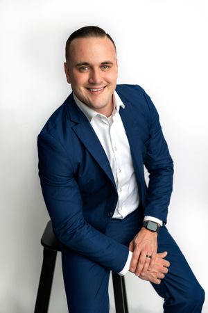 Smiling man in blue suit and white shirt sitting on stool, professional headshot on white background.