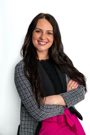 Smiling woman with dark hair in a tweed blazer, black top, and pink skirt, posing with arms crossed.
