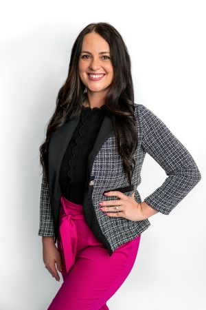 Professional woman smiling in pink pants, tweed blazer, and black blouse poses confidently against white background.