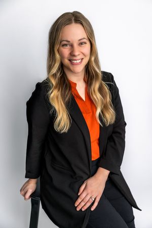 Smiling woman with long blonde hair wearing a black blazer and orange blouse, seated against a white background.