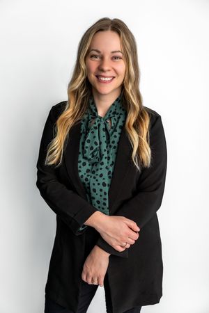 Smiling woman with blonde hair wearing a black blazer and green polka dot blouse poses against white background.
