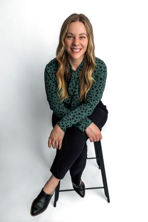 Smiling woman with long blonde hair sits on a black stool wearing a green polka dot blouse and black pants.